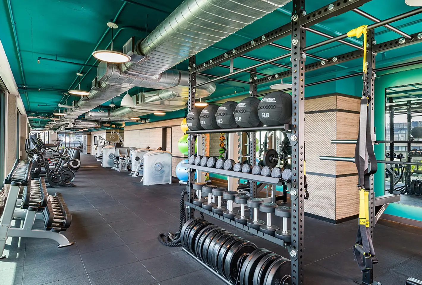 Industrial-style fitness center with weightlifting equipment, vibrant teal ceiling, and exposed air ducts