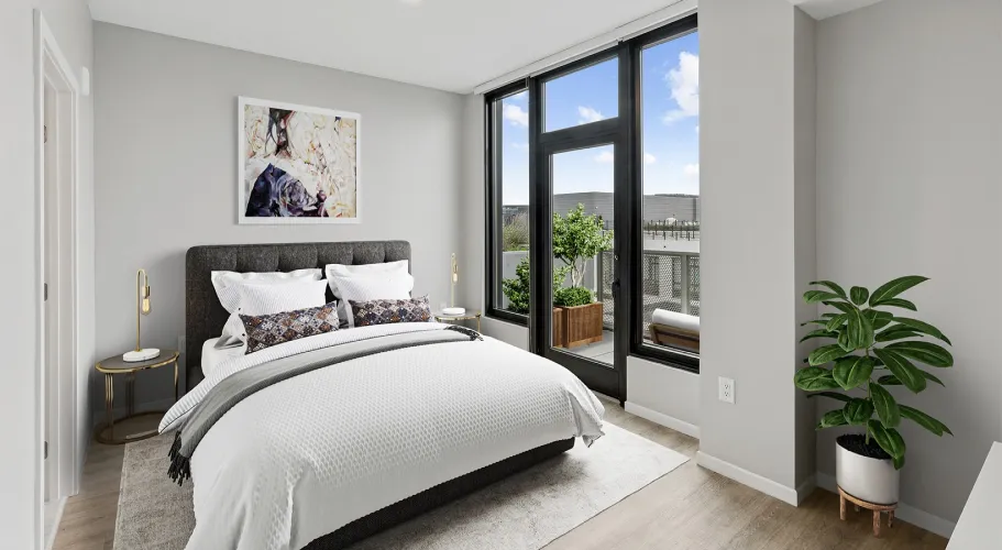 Bright bedroom with white bed, stylish desk, and large windows offering urban views