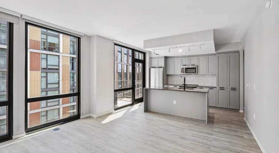 Empty modern apartment with open kitchen, large windows, and light wood flooring