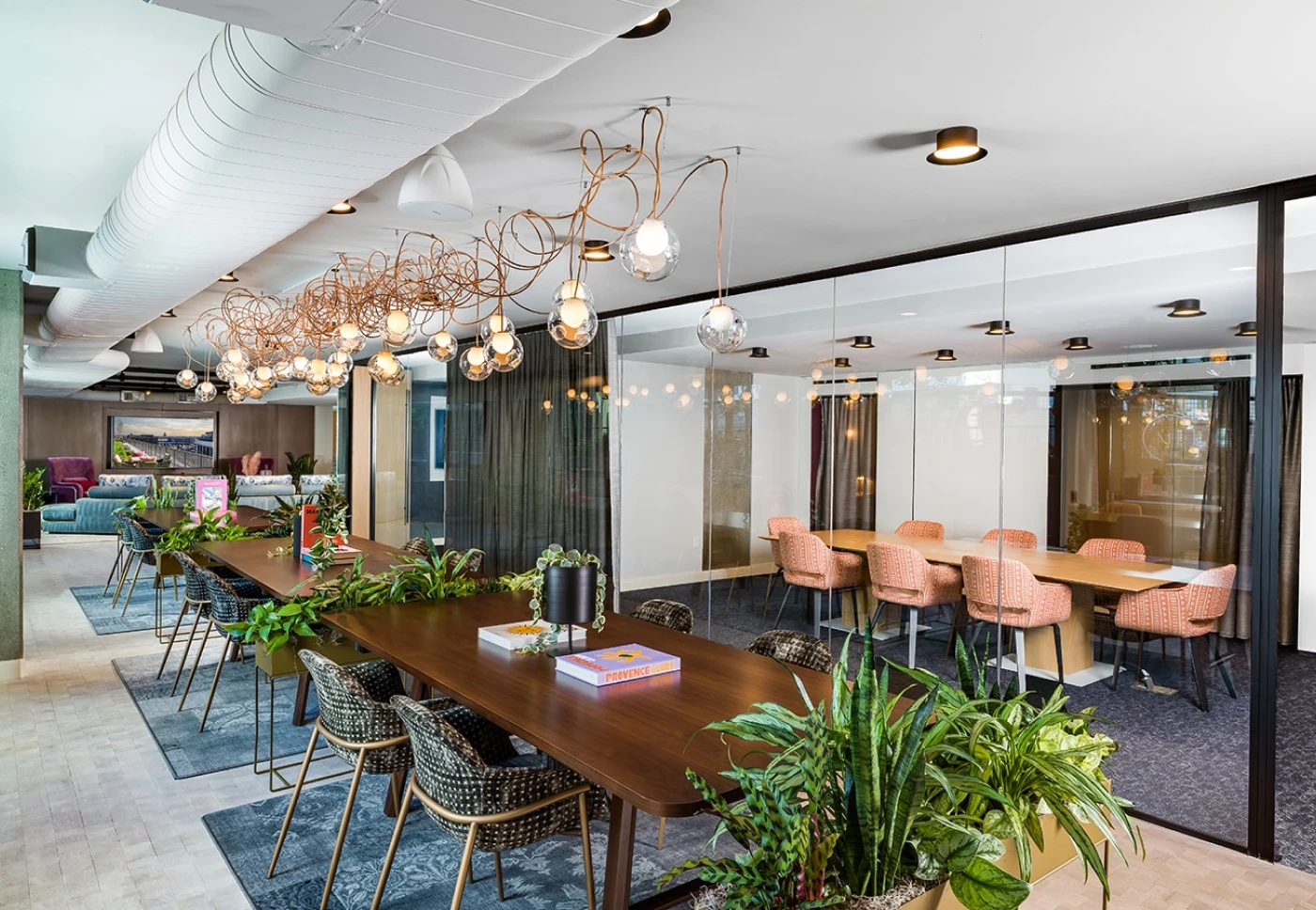 Modern coworking space with long wooden tables, lush greenery, and artistic pendant lighting